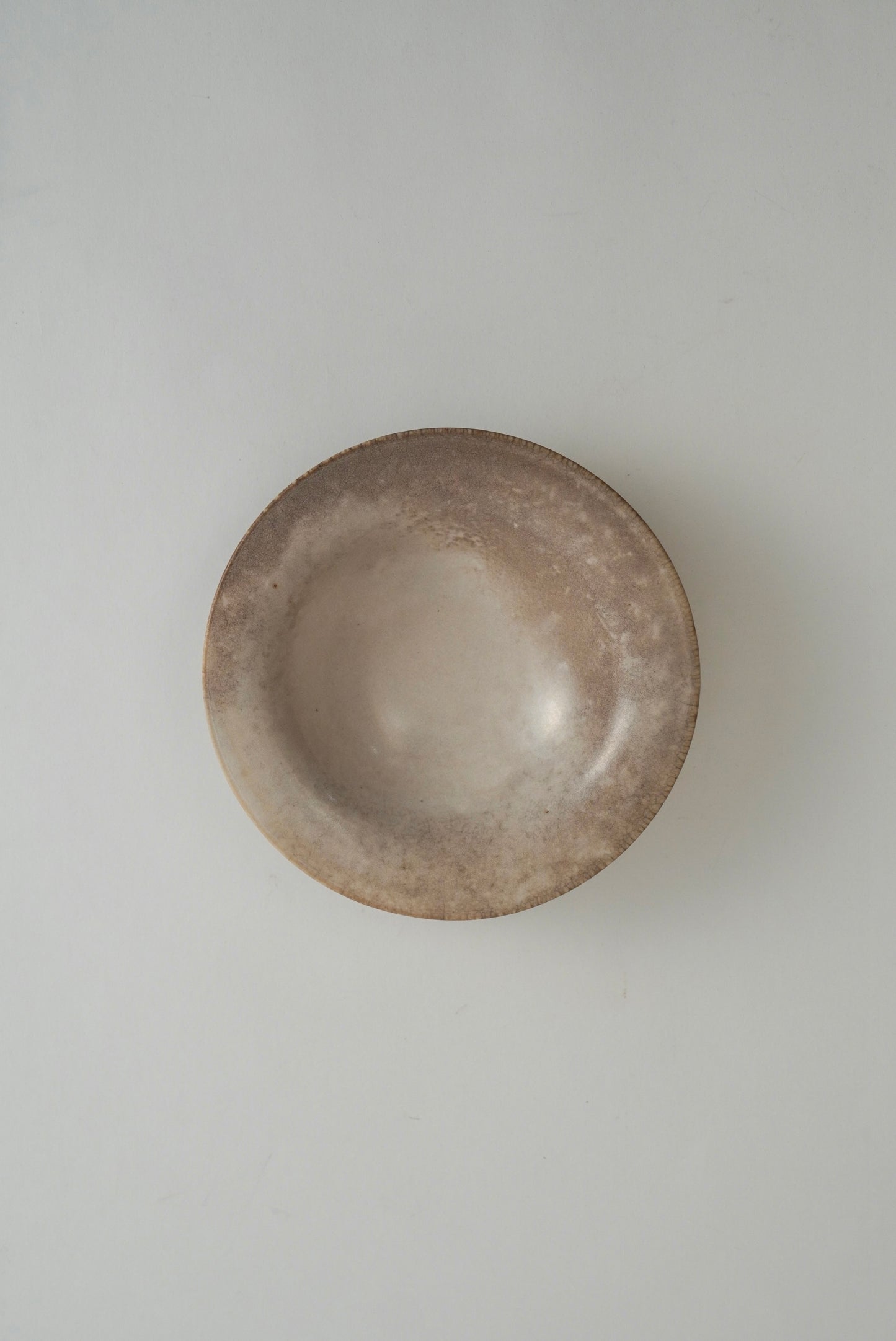 A small, round ceramic bowl with a rustic, textured finish in earthy brown and beige tones, photographed from directly above on a neutral light gray background.