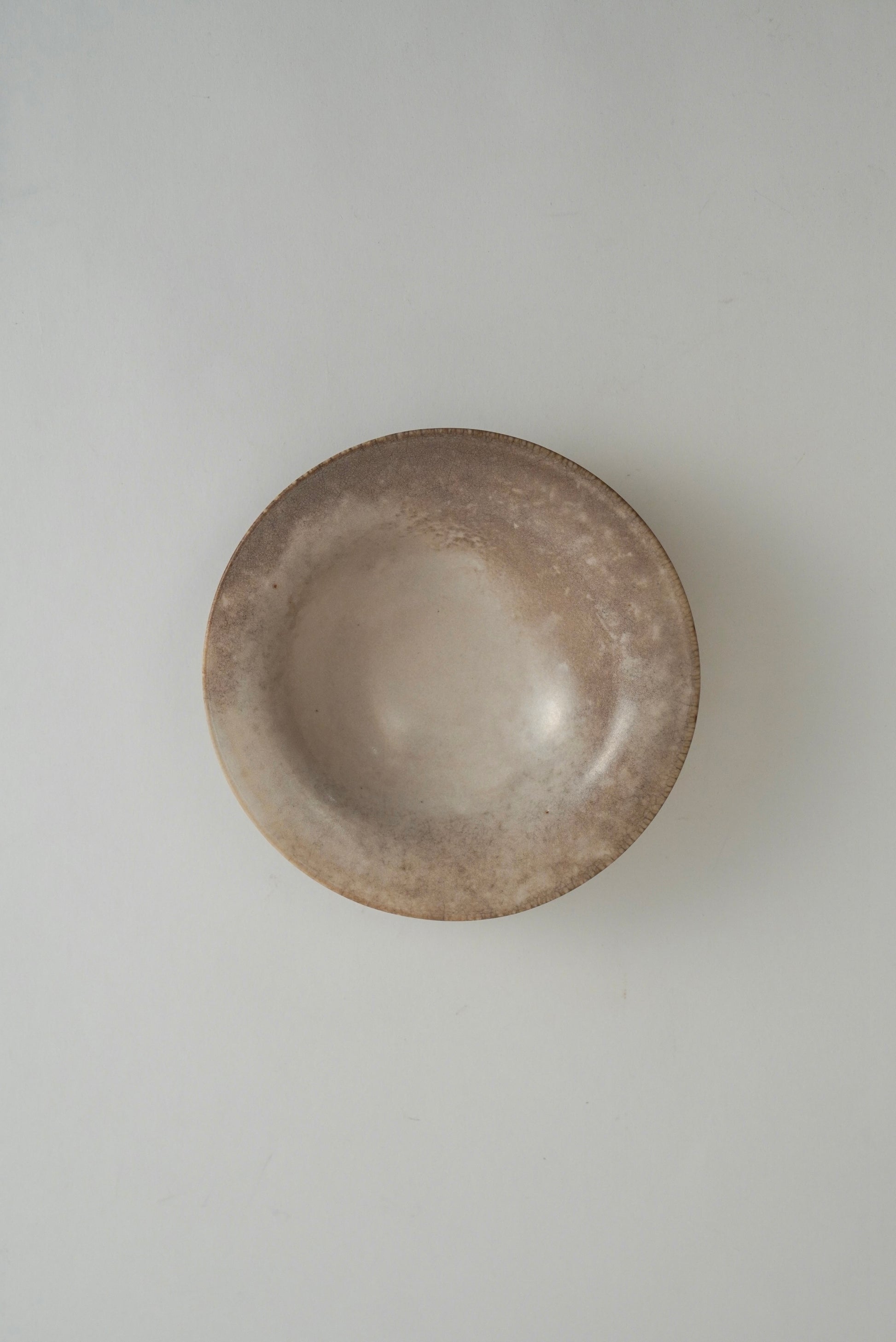 A small, round ceramic bowl with a rustic, textured finish in earthy brown and beige tones, photographed from directly above on a neutral light gray background.
