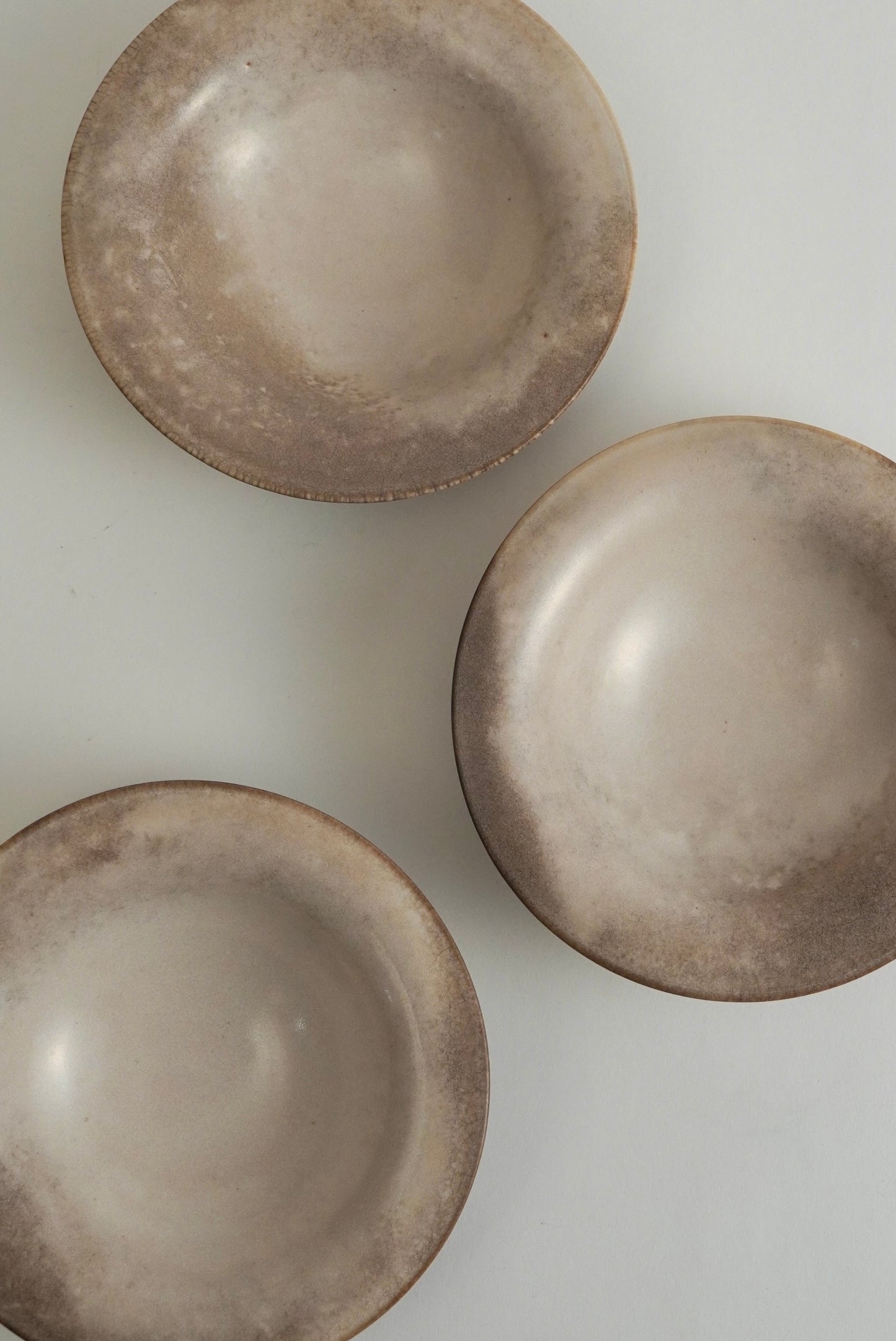 Three shallow round ceramic bowls with a textured, rustic matte finish in warm taupe and beige tones, arranged on a light off-white background.