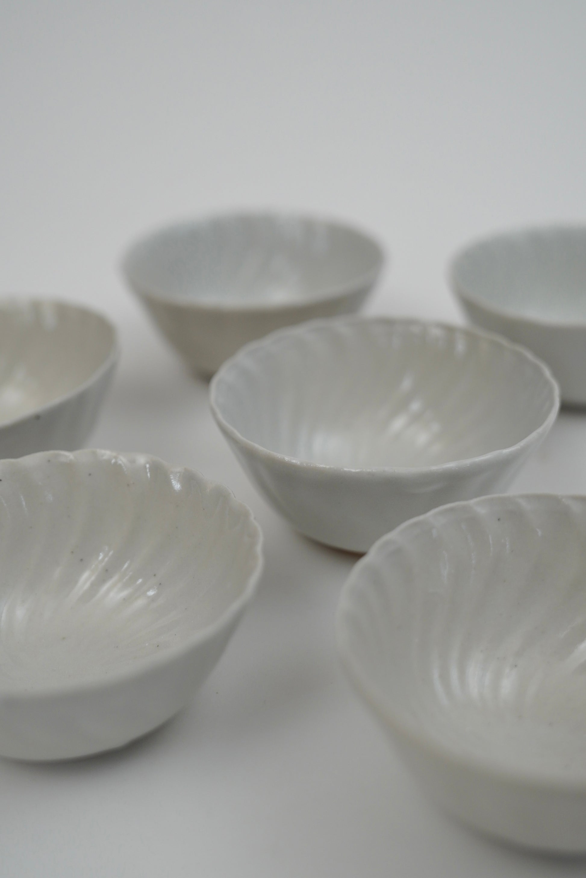 Six small white ceramic bowls with a ribbed, scalloped rim, arranged on a plain light gray background.