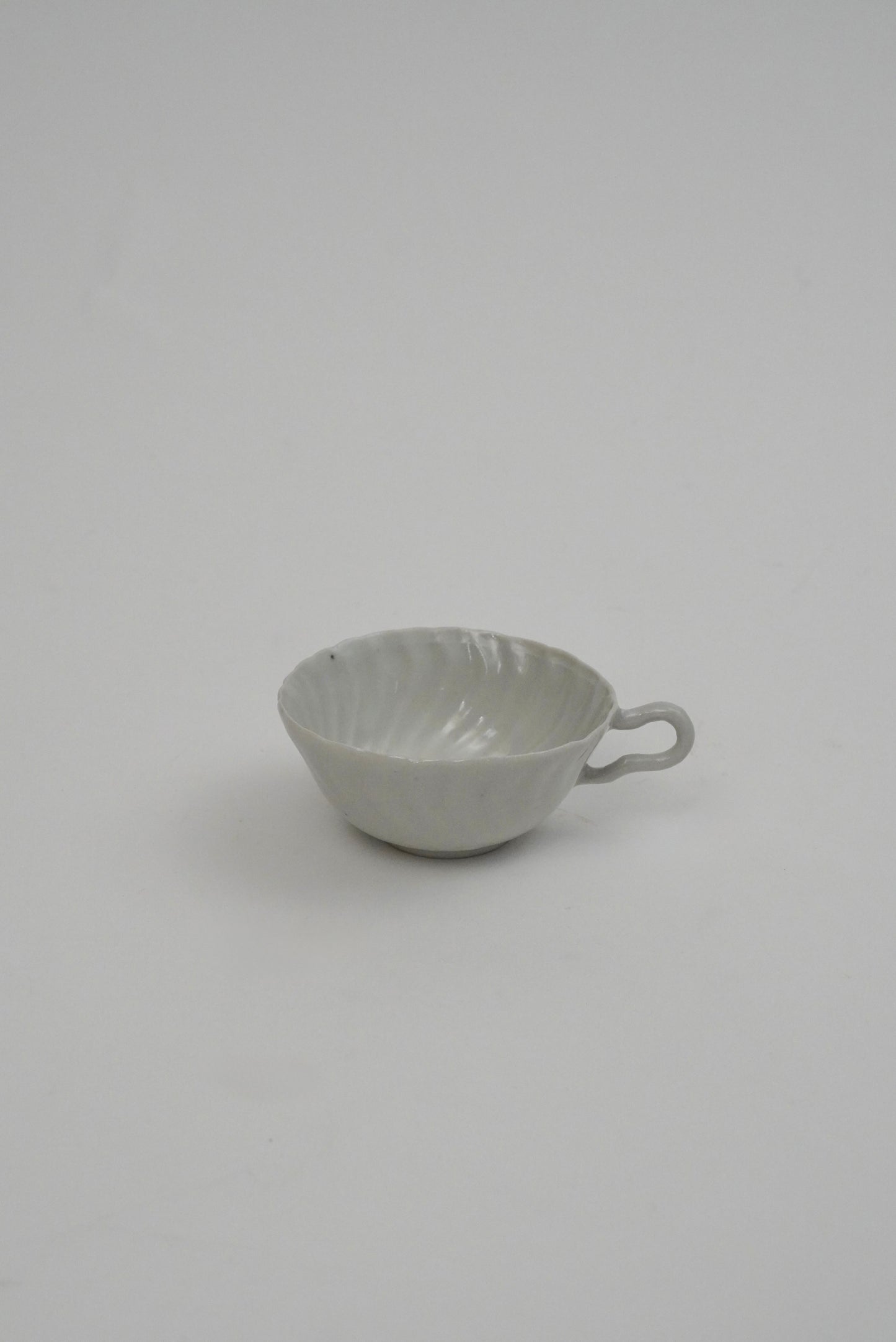 A small white ceramic cup with a ribbed exterior and a loop handle, photographed against a plain light gray background.