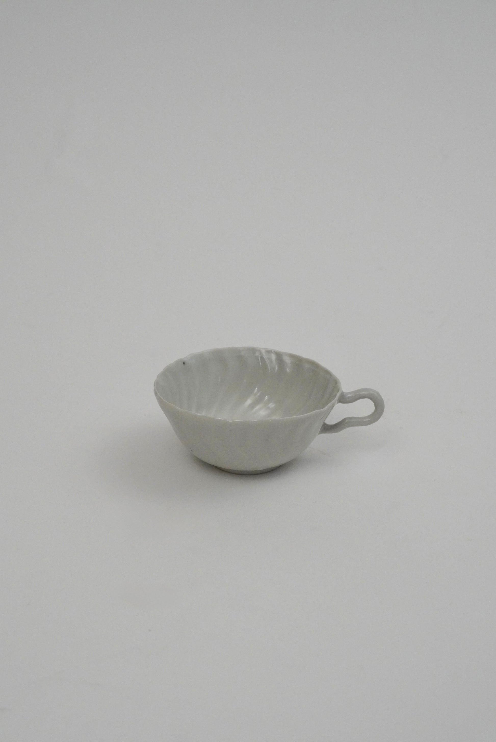 A small white ceramic cup with a ribbed exterior and a loop handle, photographed against a plain light gray background.