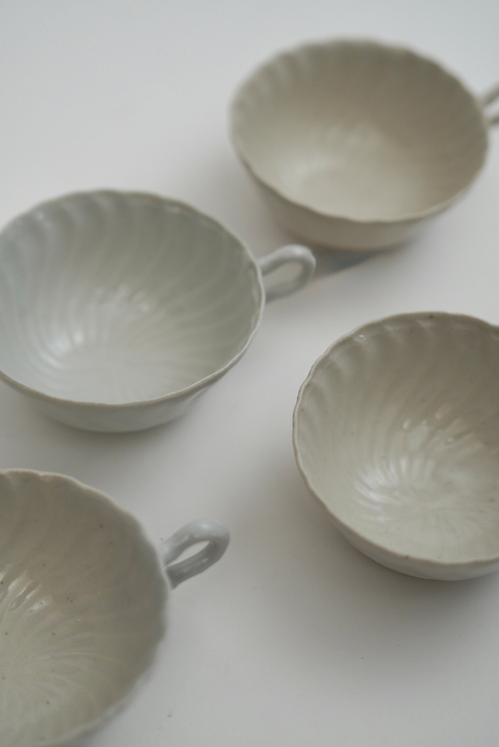 Four small ceramic bowls with scalloped rims and loop handles, shown in a soft off-white glaze on a plain light background.