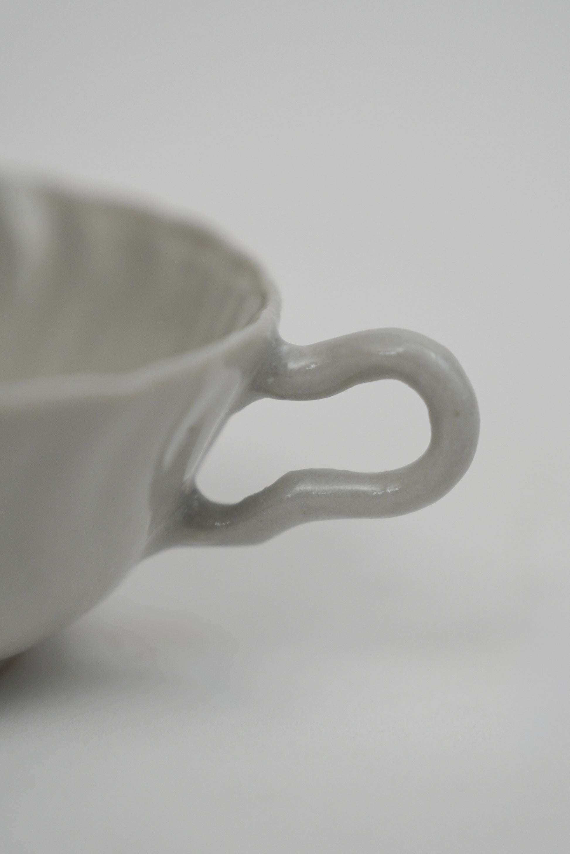 Close-up of a light gray ceramic cup with a wavy, looped handle on a plain light gray background.
