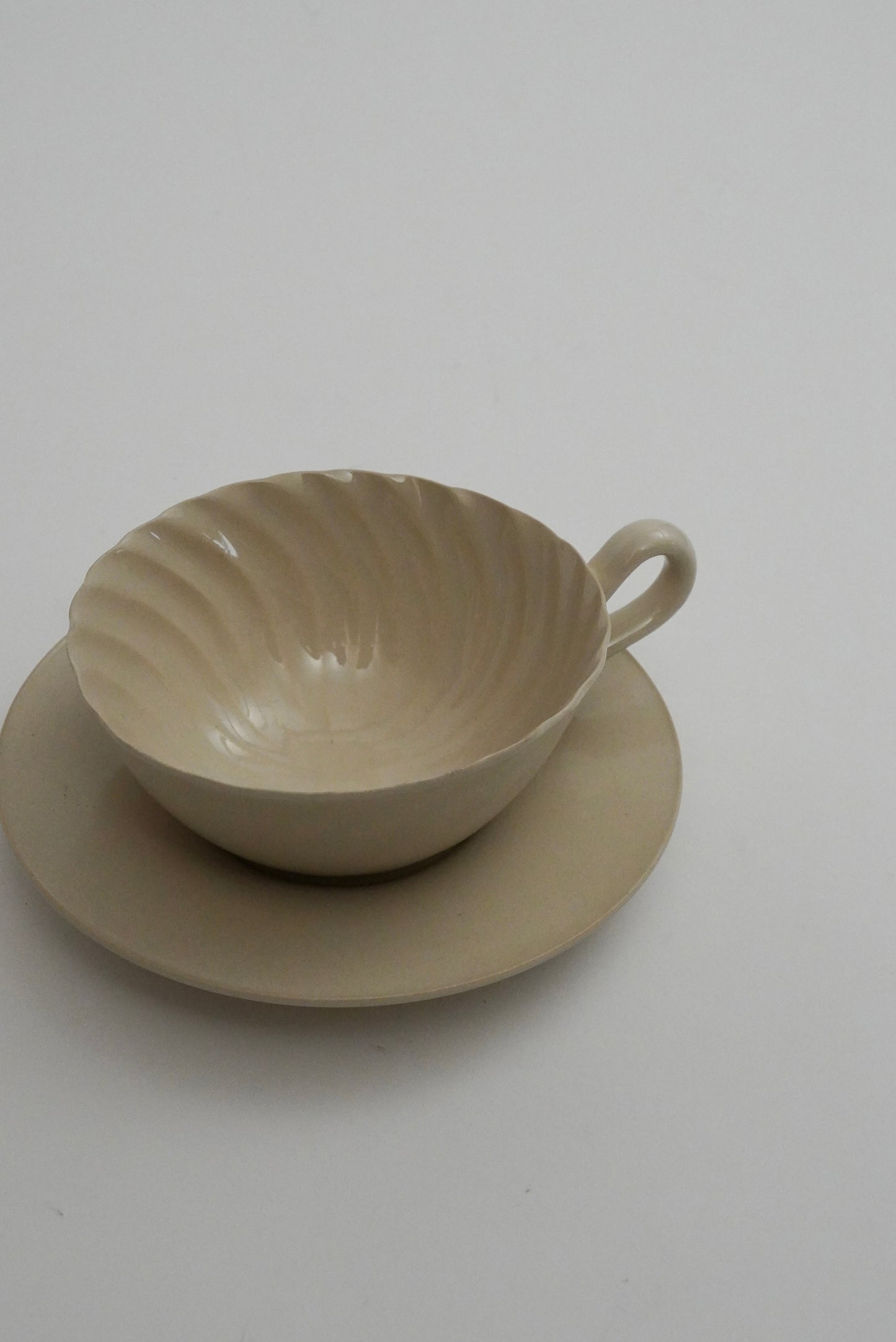A light beige ceramic teacup with a fluted, ribbed interior and a matching saucer, photographed on a plain light gray background.