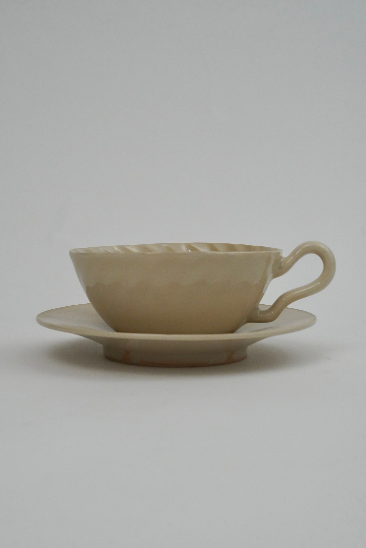 A beige ceramic teacup with a matching saucer, featuring a textured, hammered surface and a curved handle, set against a plain light gray background.