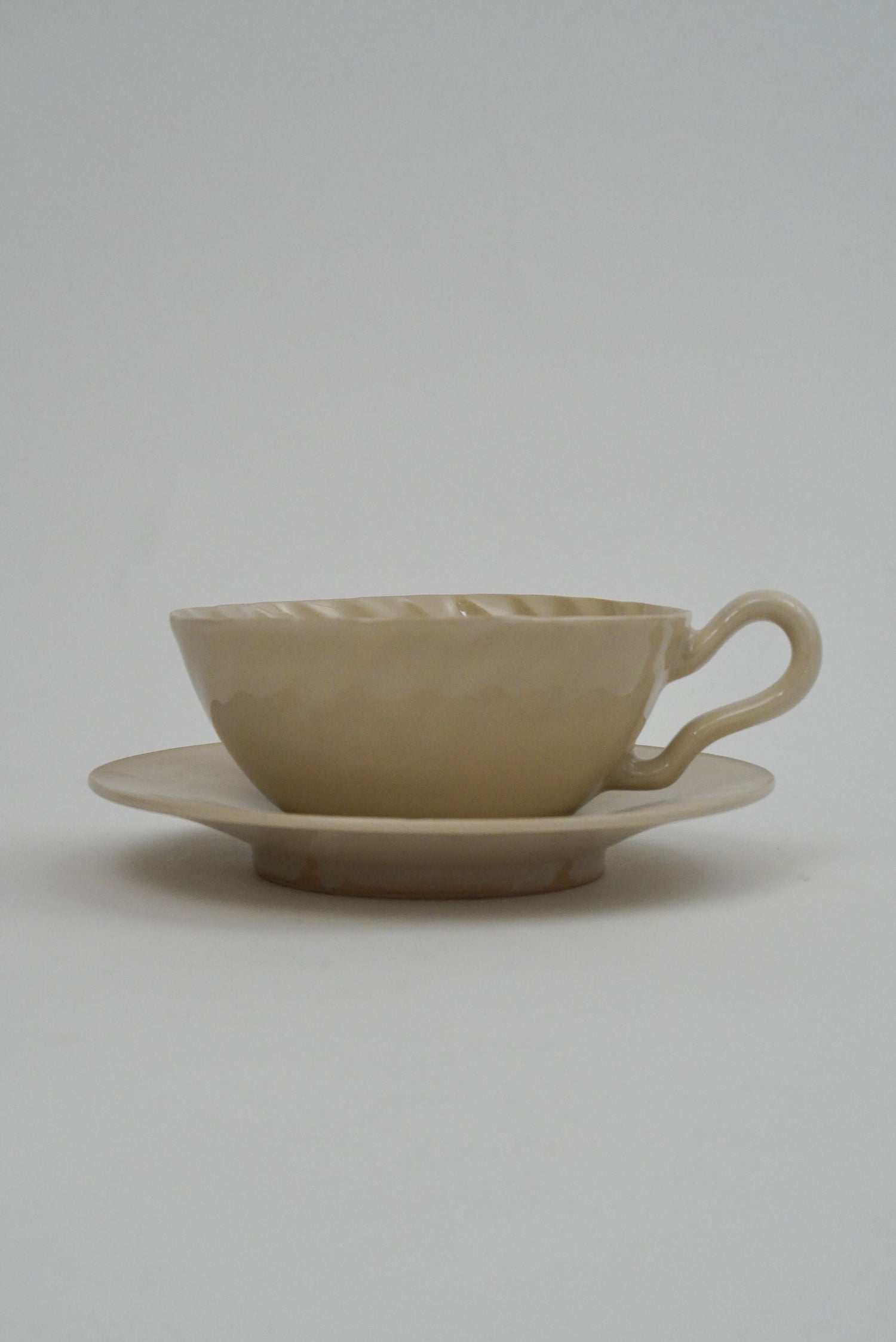 A beige ceramic teacup with a matching saucer, featuring a textured, hammered surface and a curved handle, set against a plain light gray background.