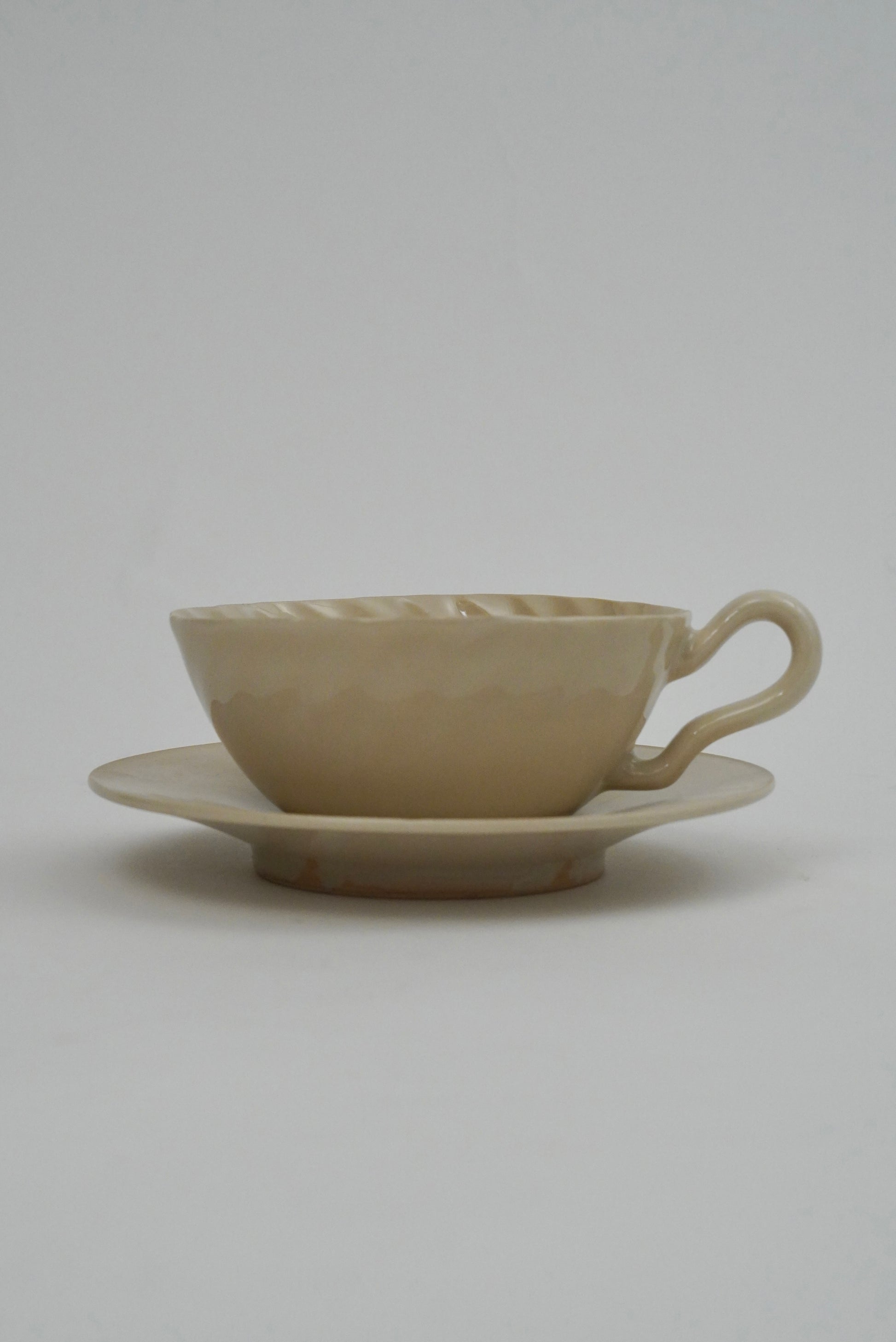 A beige ceramic teacup with a matching saucer, featuring a textured, hammered surface and a curved handle, set against a plain light gray background.