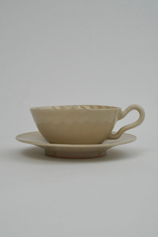 A beige ceramic teacup with a matching saucer, featuring a textured, hammered surface and a curved handle, set against a plain light gray background.