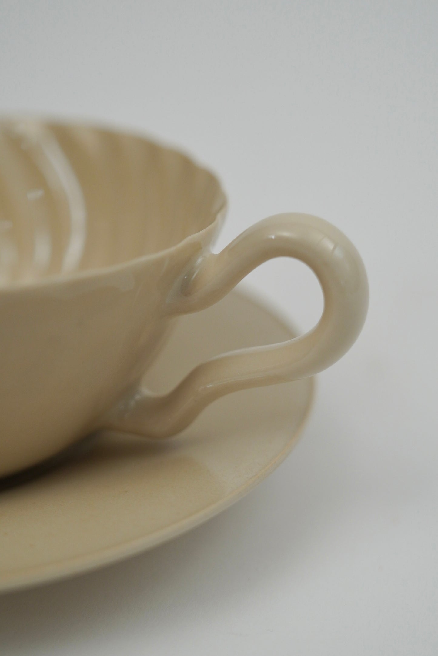 A close-up of a beige ceramic teacup with a matching saucer, featuring a wavy handle and ribbed interior, set against a plain light gray background.