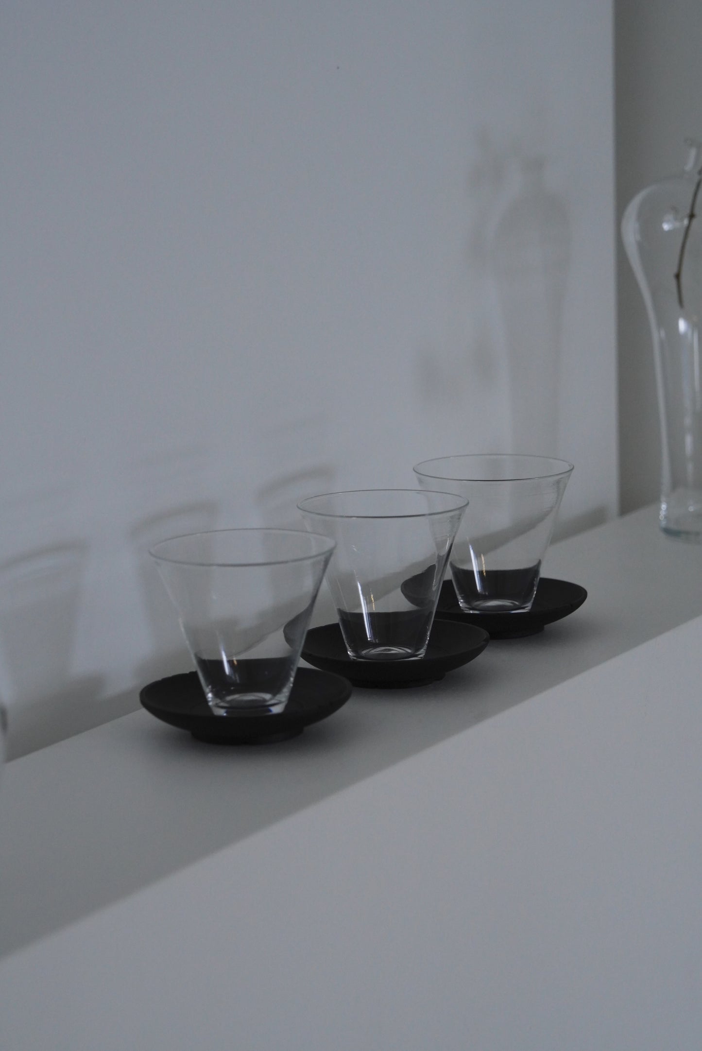 Three clear glass tumblers on black round trays against a white wall. Handmade wooden saucer by Japanese artist at Gallery tete Hong Kong.