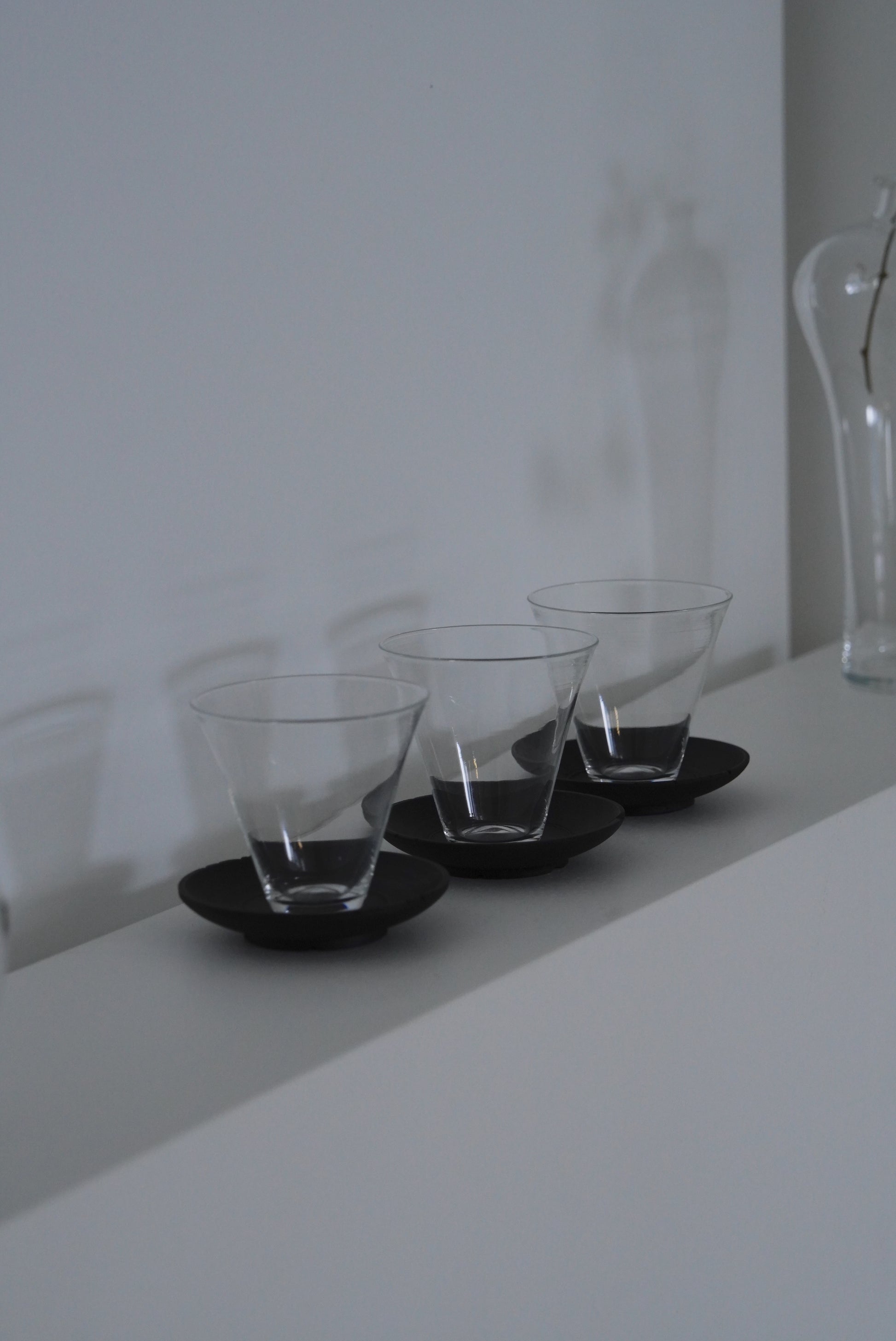 Three clear glass tumblers on black round trays against a white wall. Handmade wooden saucer by Japanese artist at Gallery tete Hong Kong.