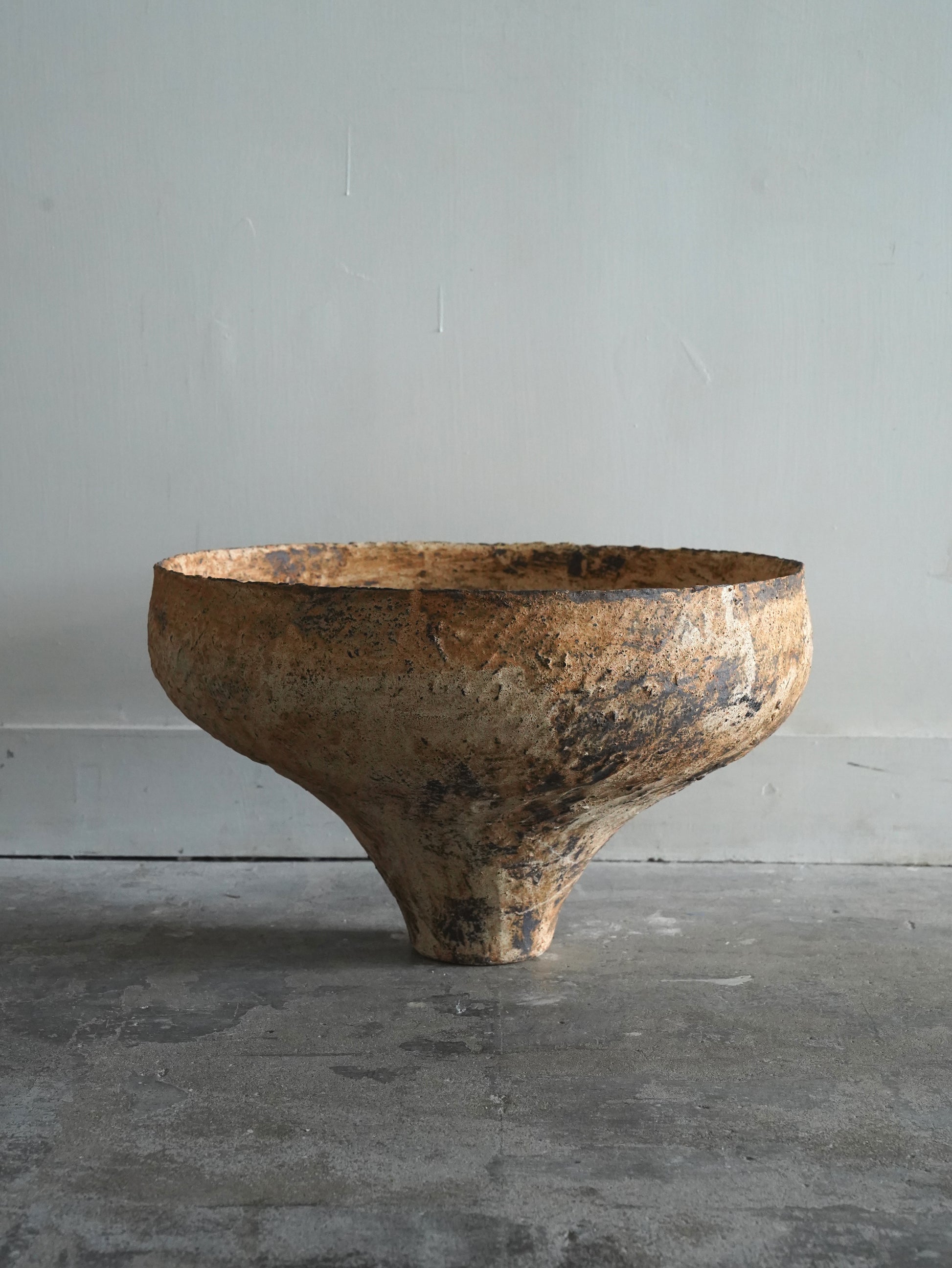 A rustic, handcrafted clay bowl with a wide, shallow top and a tapered base, shown on a concrete floor against a plain gray wall.