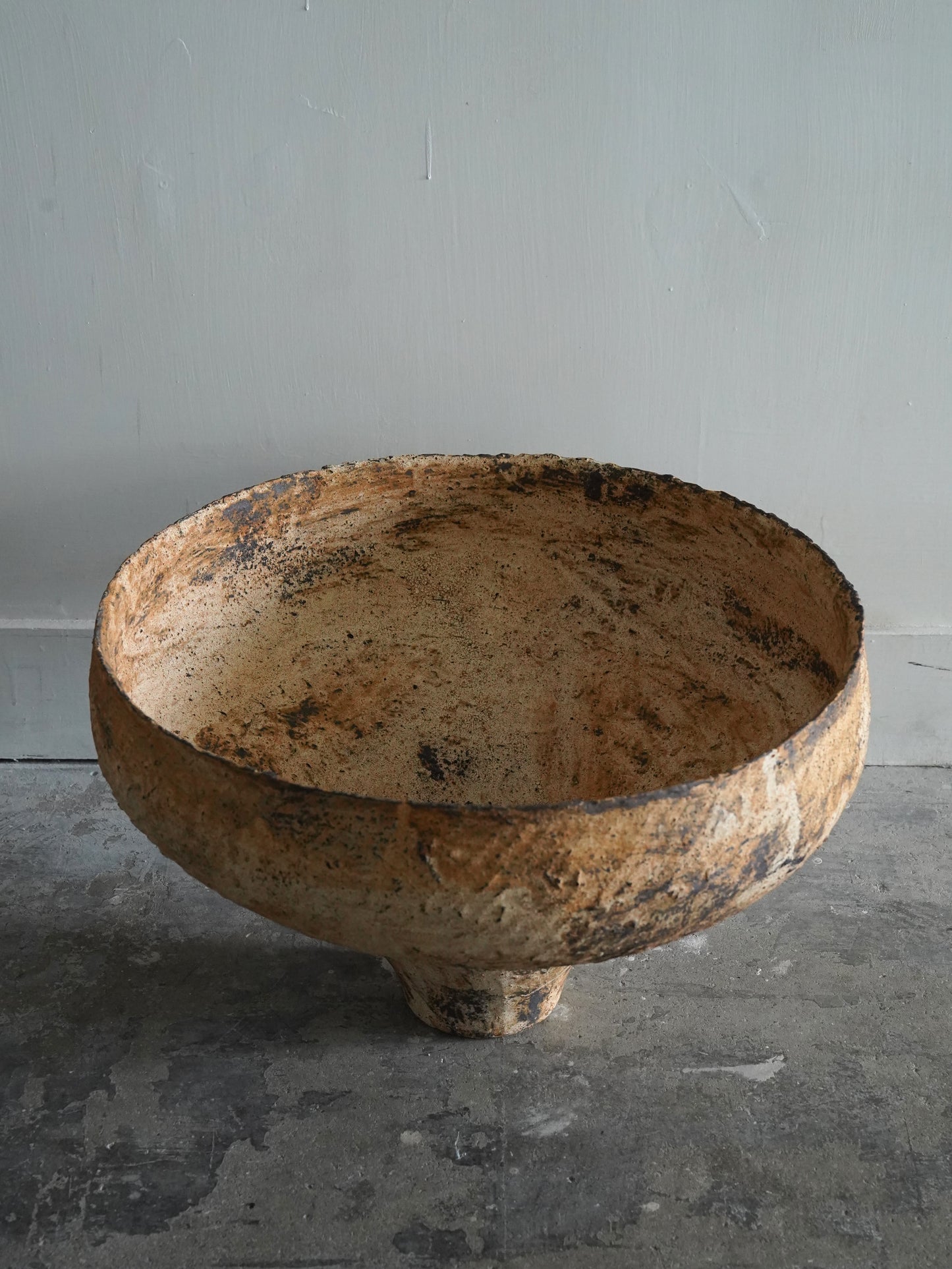 A large, rustic ceramic bowl with a textured, distressed finish in earthy brown and beige tones, set against a neutral gray wall and concrete floor.