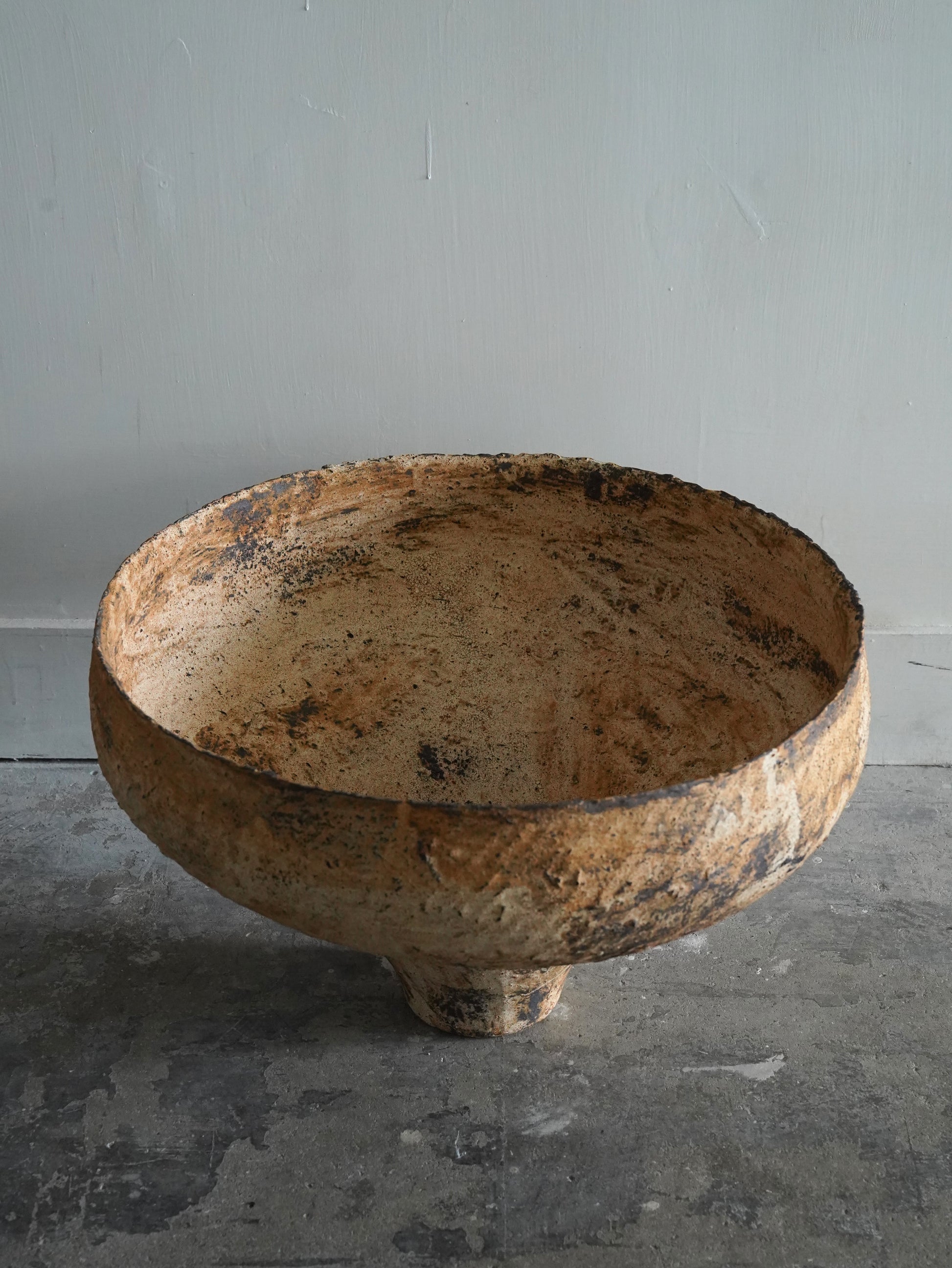 A large, rustic ceramic bowl with a textured, distressed finish in earthy brown and beige tones, set against a neutral gray wall and concrete floor.