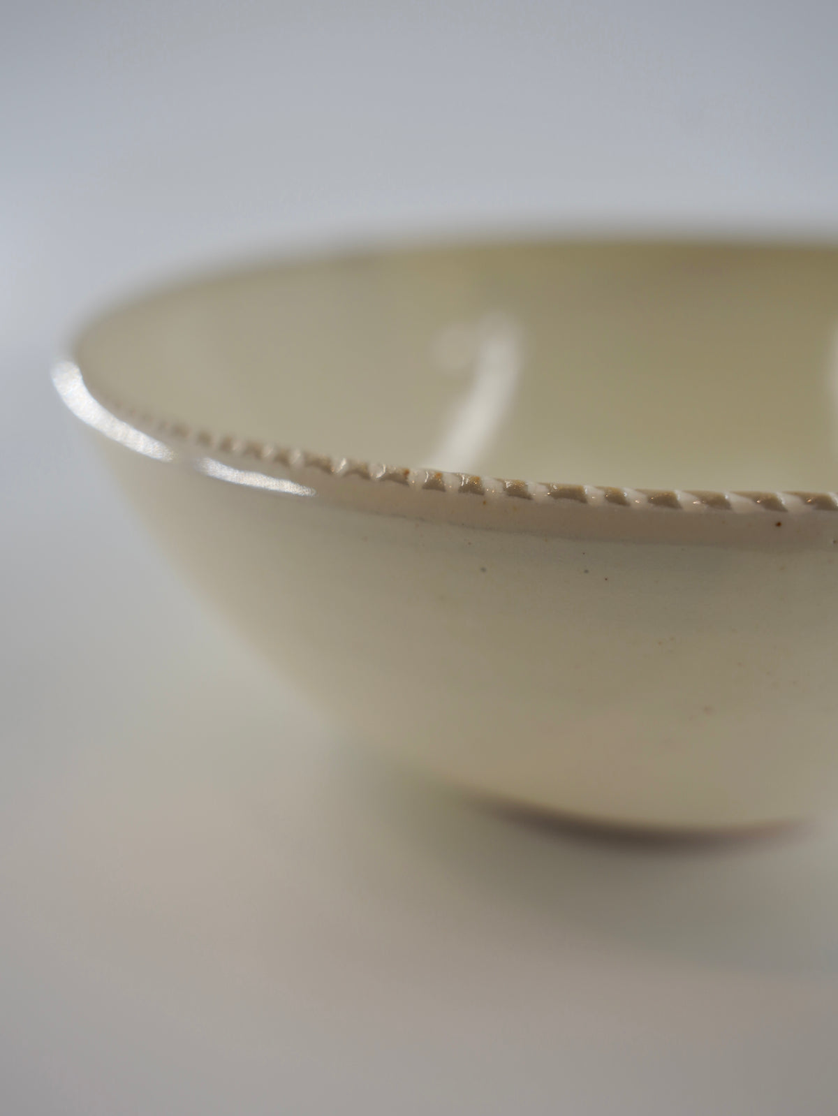 Close-up of a ceramic bowl with a glossy cream-colored interior and a textured, hand-painted beige rim, shown against a neutral light gray background.