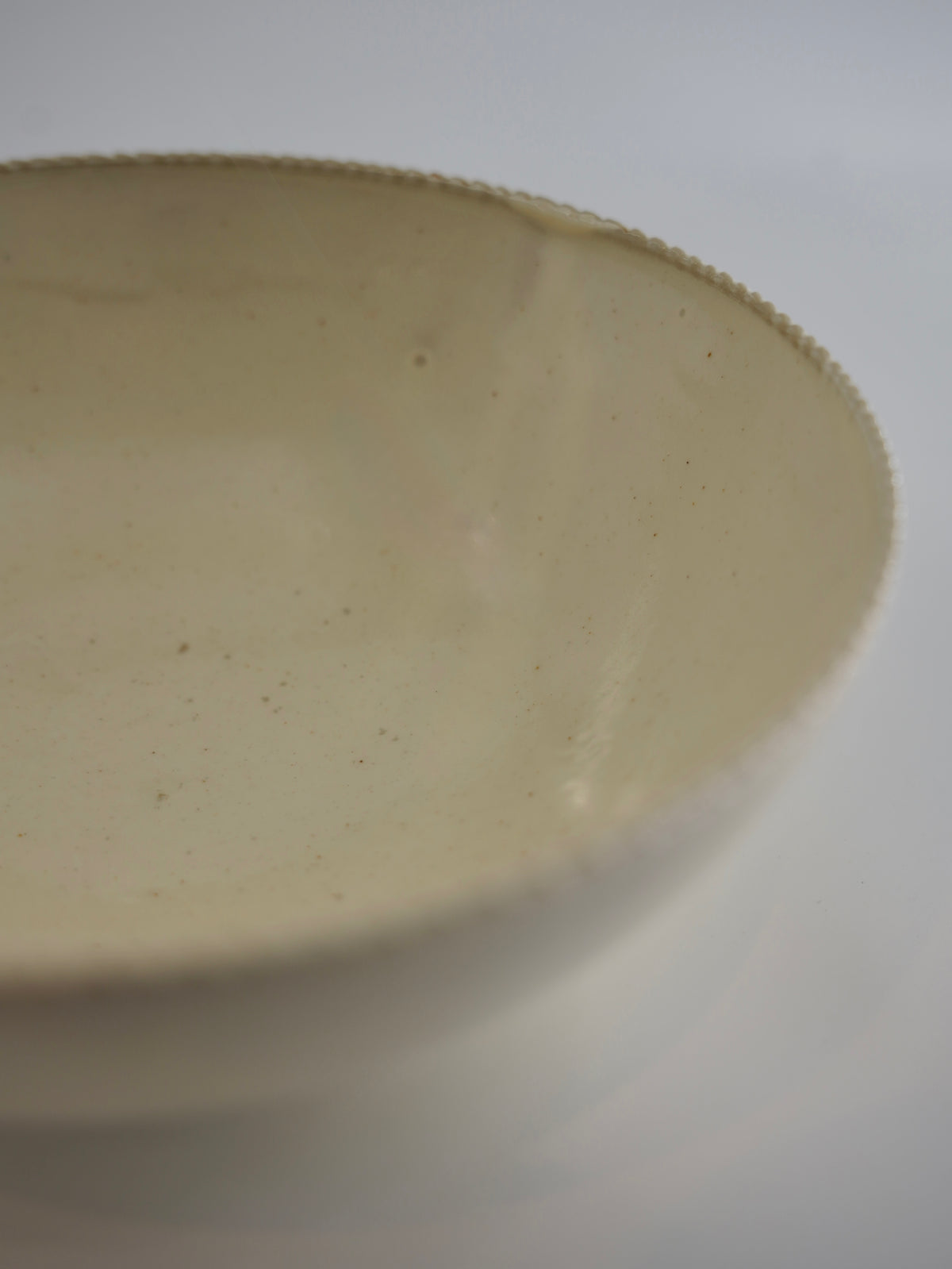 A close-up of a ceramic bowl with a warm cream-beige glaze and subtle speckles, showing the textured rim against a neutral light gray background.