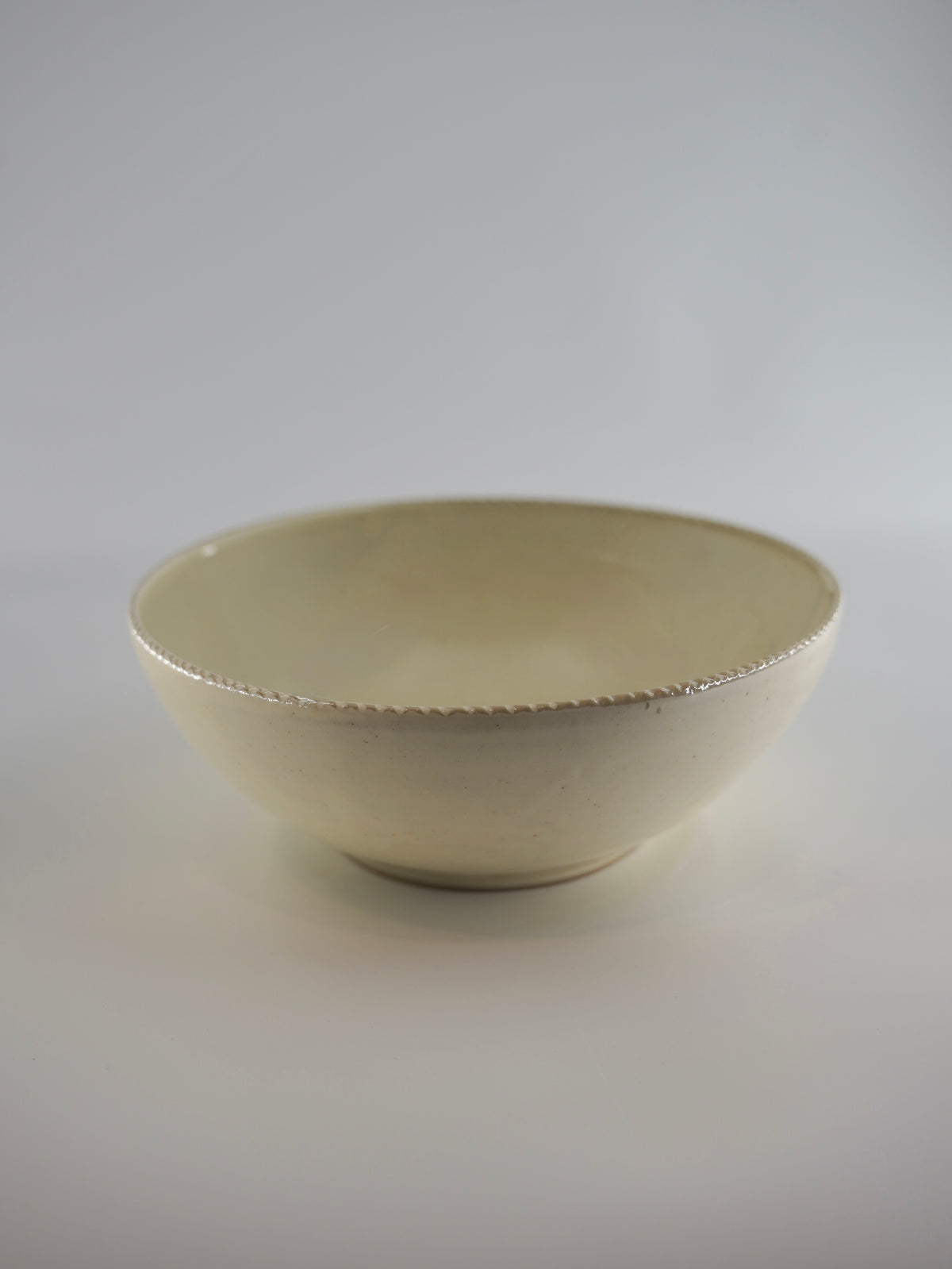 A handmade ceramic bowl with a textured, scalloped rim in a warm cream or off-white glaze, photographed against a neutral light gray studio background.