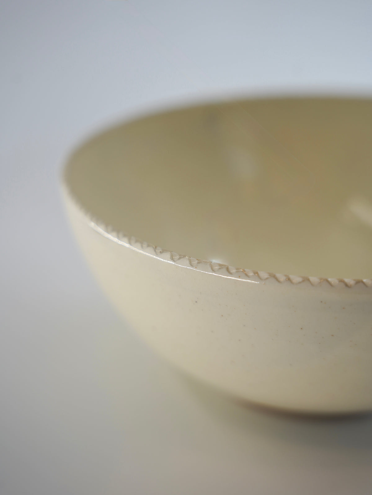 A close-up of a ceramic bowl with a warm cream-colored glaze and a delicate, textured rim, shown against a neutral light gray background.