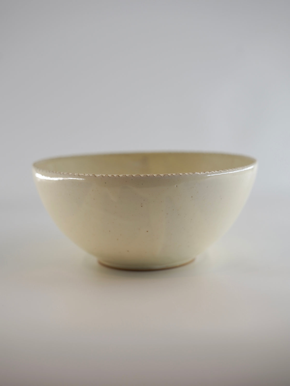A handmade ceramic bowl with a matte, warm off-white glaze and a subtle scalloped rim, photographed against a neutral light gray background.